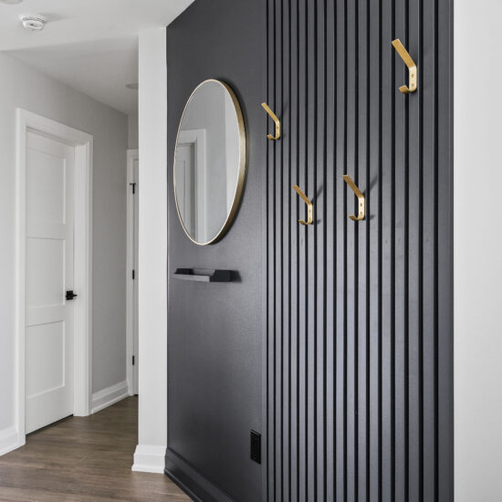 Stylish entryway in a Whitby home featuring a black slatted accent wall, gold hooks, a round mirror, and wood flooring for a sleek modern look.