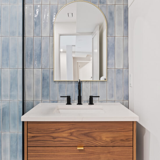 Floating wood vanity with a white quartz countertop, matte black faucet, and arched mirror against glossy blue vertical subway tiles in a modern Durham Region bathroom.