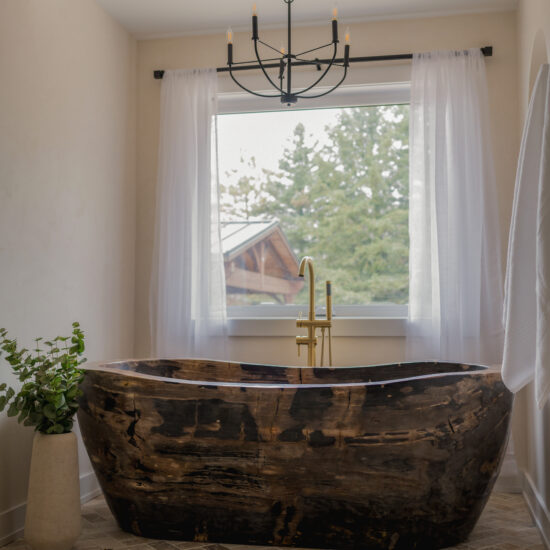 Elegant bathroom featuring a handcrafted wooden bathtub, gold fixtures, a black chandelier, and natural light from a large window with sheer curtains, designed by MM Interior Design for homes in Port Perry and Uxbridge.