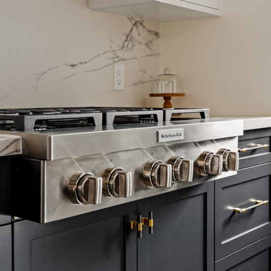Close-up of a high-end stainless steel KitchenAid range set against a sleek marble backsplash with dark cabinetry and gold-accented hardware, designed by MM Interior Design for modern kitchens in Whitby, Oshawa, and Clarington.