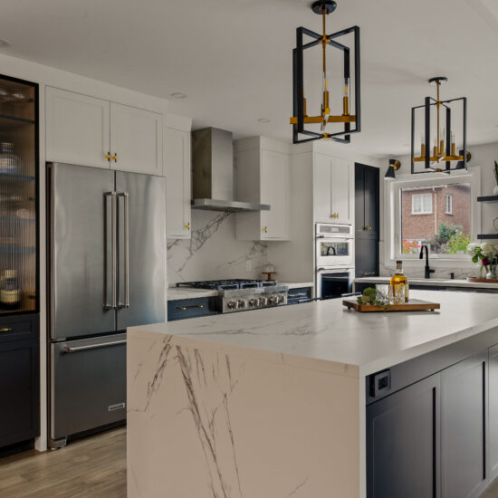 Spacious modern kitchen featuring a large marble waterfall island, two-tone cabinetry in white and charcoal, gold-accented pendant lighting, and high-end stainless steel appliances, designed by MM Interior Design for homes in Whitby, Oshawa, and Uxbridge.