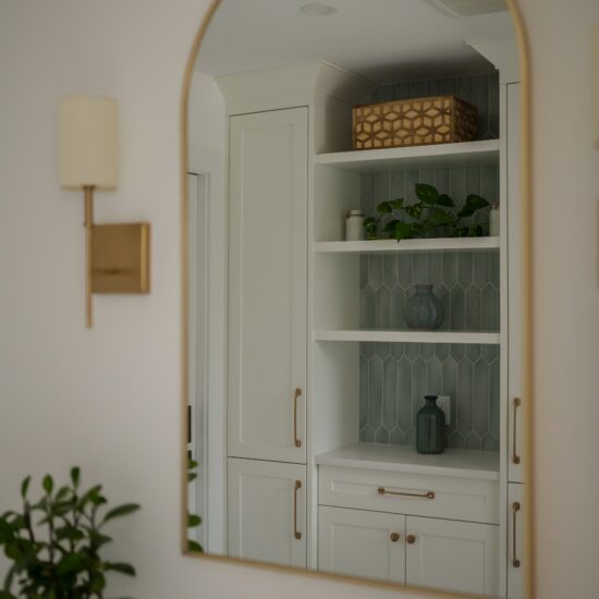 A stylish bathroom featuring an arched mirror reflecting built-in white cabinetry with brass hardware and open shelving. The shelves are accented with green plants, decorative vases, and a geometric-patterned backsplash, creating a sophisticated and modern design