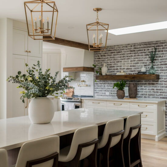 A modern farmhouse kitchen featuring a long island with white countertops, cushioned barstools with wooden frames, gold lantern pendant lighting, and a white brick backsplash with wooden floating shelves.
