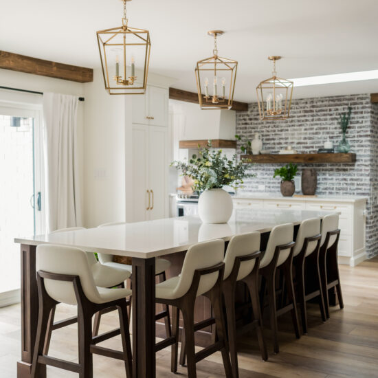 A spacious modern farmhouse kitchen featuring a large island with wooden barstools and white cushions, gold geometric pendant lighting, and a white brick backsplash with floating wooden shelves.