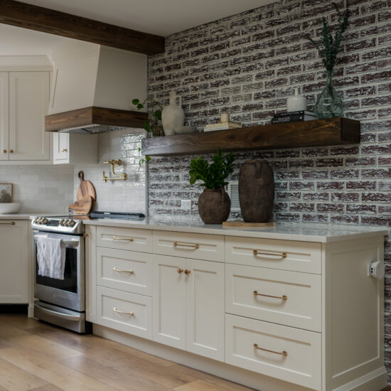 A modern farmhouse kitchen with white cabinetry, gold hardware, a rustic white brick backsplash, and dark wood floating shelves adorned with vases and greenery. The kitchen also features a farmhouse-style range hood with wood trim and a brass pot filler above the stove.