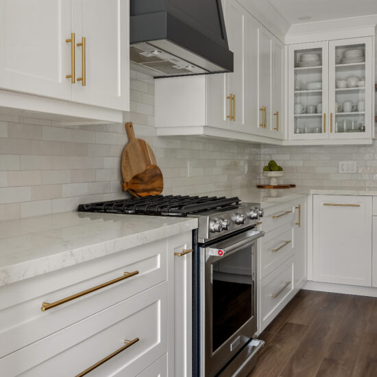 Modern kitchen featuring white shaker cabinets, a black range hood, gold hardware, and a stainless steel gas range with a marble countertop