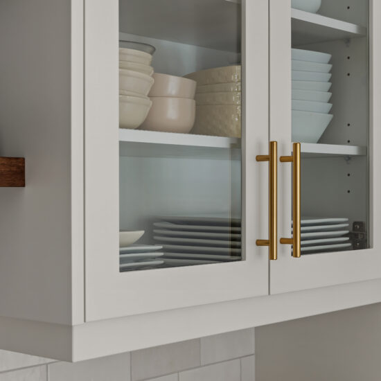 Close-up of white glass-front kitchen cabinets with gold handles, showcasing neatly stacked dishes and bowls, alongside a wooden floating shelf with cookbooks.