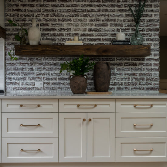 A modern farmhouse kitchen with white shaker cabinets, gold hardware, a rustic whitewashed brick backsplash, and a floating dark wood shelf decorated with ceramic vases, books, and greener