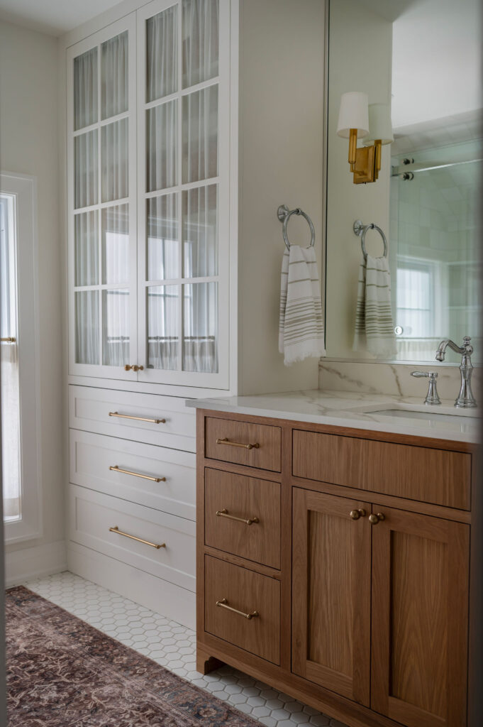 A view of a Whitby bathroom showing a floor-to-ceiling white built-in linen cabinet with glass doors and fabric inserts. Next to it is a light oak vanity with a marble top, brass faucet, and a brass wall sconce.