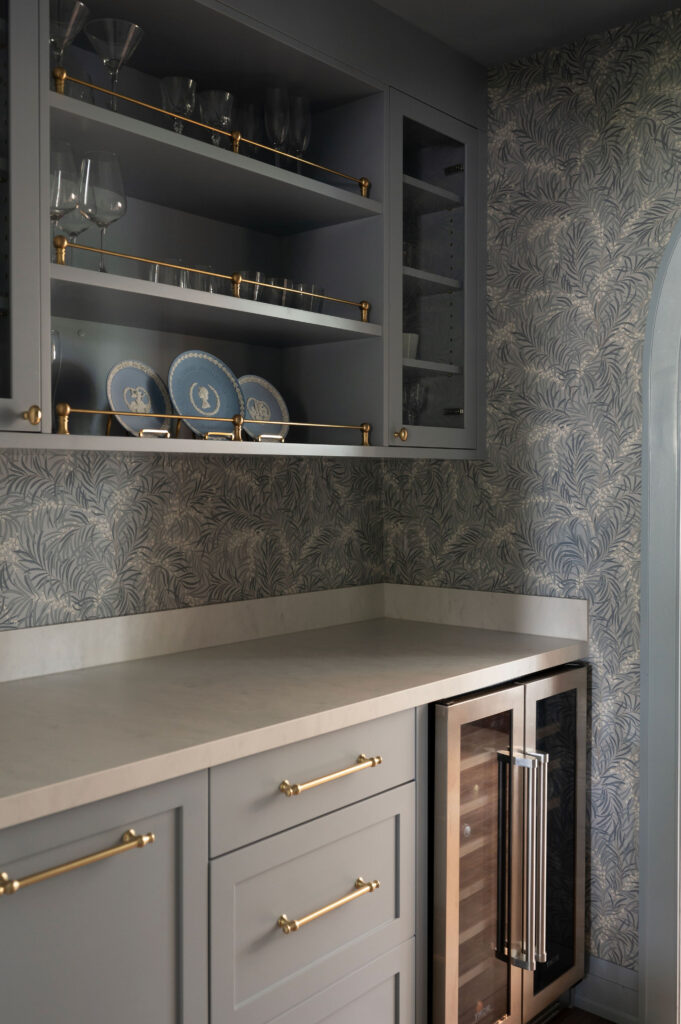 A butler's pantry in Uxbridge with dusty blue cabinetry, brass hardware, a built-in wine fridge, and patterned blue and grey wallpaper.