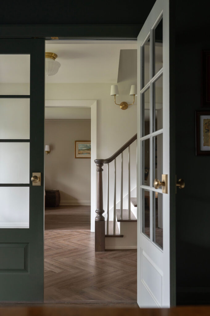 View through dark green French doors into a hallway in an Uxbridge home featuring light oak herringbone hardwood floors and a white staircase with a dark wood handrail.
