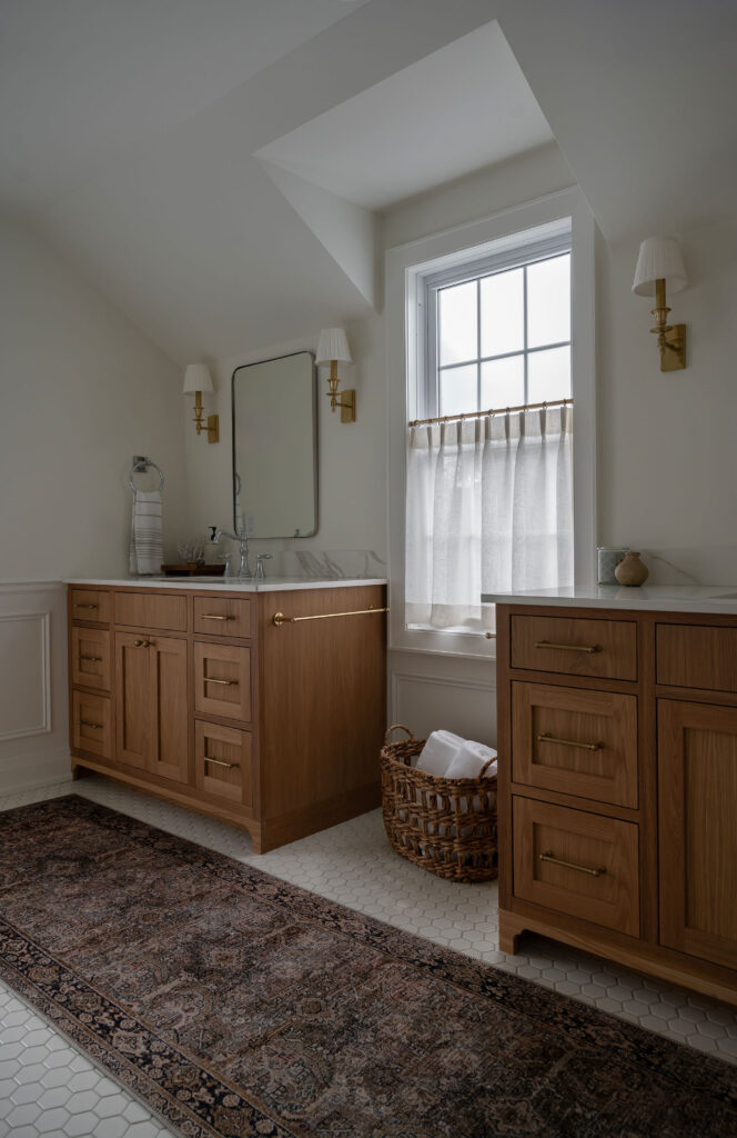 A bright Whitby bathroom featuring two custom light-oak vanities with gold hardware. A long vintage-style patterned runner sits on a white hexagon tile floor between the vanities, and brass sconces flank a minimalist mirror.