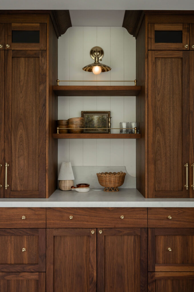 Custom walnut wood cabinetry in an Uxbridge home bar featuring open shelving with brass gallery rails and a vintage-style brass wall sconce.