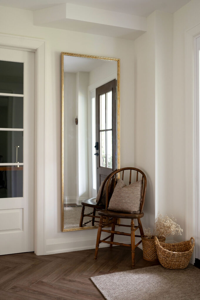 A tall gold-framed floor mirror leaning against a white wall in an Uxbridge entryway, next to a vintage wooden chair and woven baskets on herringbone floors.