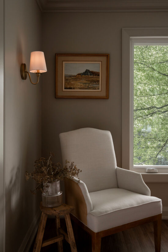 A neutral-toned reading corner in an Uxbridge home with a white high-back armchair, a brass wall sconce, and a framed vintage landscape painting.