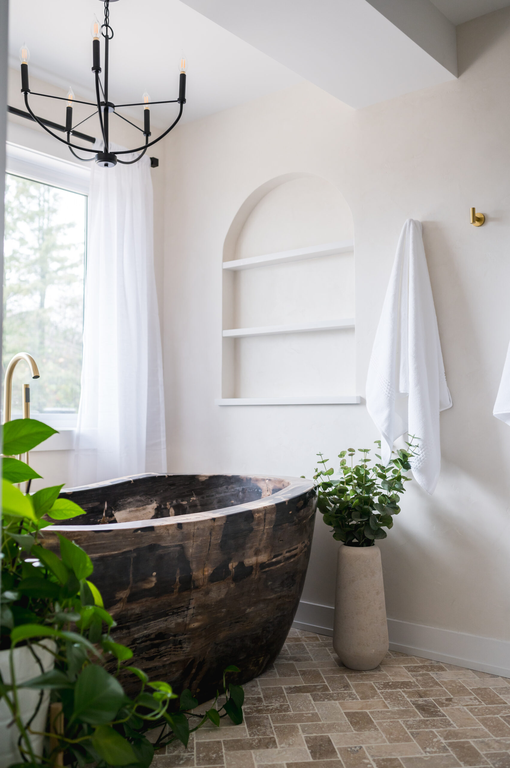 Clarington bathroom featuring a distinctive petrified wood soaker tub, herringbone travertine floor tiles, an arched wall niche, and a black cage-style chandelier.