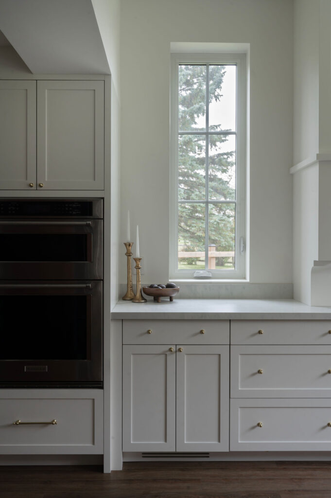 Bright white shaker-style kitchen cabinets in Uxbridge with gold hardware, a white marble countertop, and built-in double wall ovens next to a tall, narrow window.